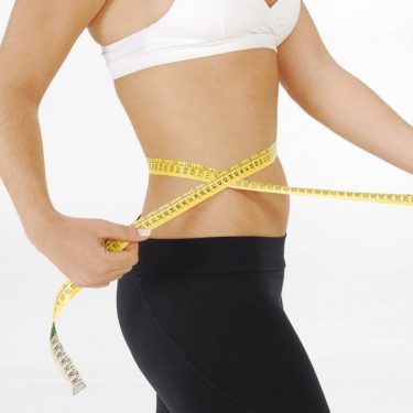 Fit woman in sportswear measuring her waist with a tape measure against a white background.