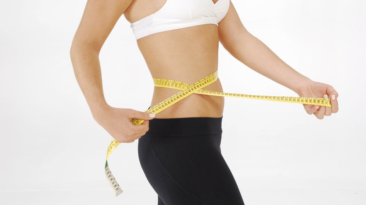 Fit woman in sportswear measuring her waist with a tape measure against a white background.